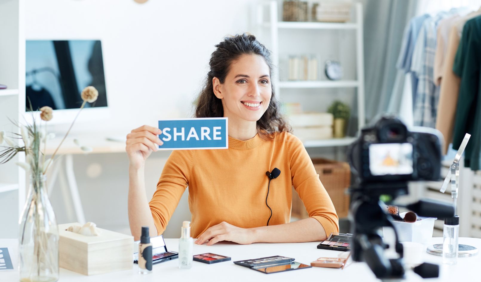 A woman blogger sitting at a table in front of a video camera holding up a sign that reads share in hopes of creating viral content.